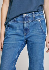 Blå denimjeans med rak passform, som har framfickor, bälteöglor och dragkedja. Tyget verkar något slitet med en smooth yta.