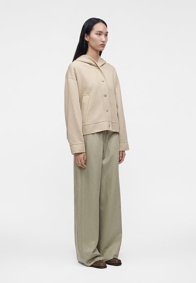 Femme portant une veste à capuche beige boutonnée, un pantalon large gris clair et des chaussures marron, debout devant un fond clair uni.