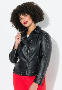 Black leather motorcycle jacket with silver studs, asymmetrical zipper, and side pockets. Worn over a white top with red pants.