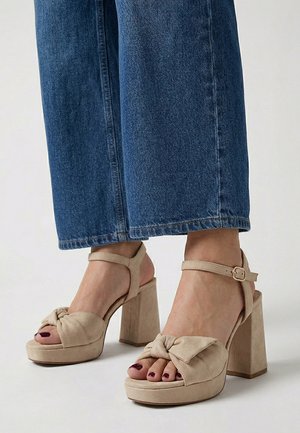 Pies con sandalias beige de tacón alto con tiras en el tobillo y diseño de nudo, combinadas con pantalones vaqueros cortos azules.