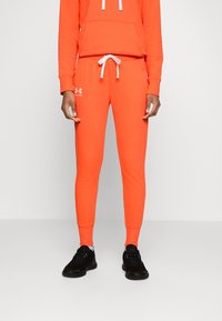 Orange träningsbyxor med dragsko i midjan, manschetter vid anklarna och en vit logotyp på vänster höft. Svarta sportskor.