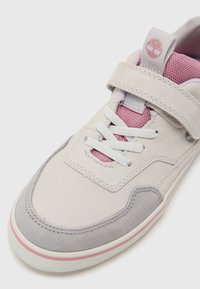 Lys grå semsket sneakers med en rosa mesh-tunge, hvite lisser og en borrelåsrem. Har en subtil rosa aksent langs sålen.