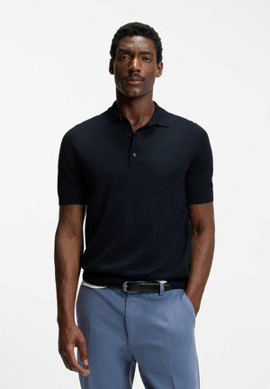 Mann mit kurzen lockigen Haaren und Schnurrbart, der ein schwarzes kurzärmeliges Poloshirt und hellblaue Hosen mit einem schwarzen Gürtel trägt.