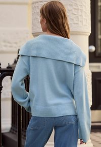 Gaiši zils džemperis ar pārlieku lielu ribota raksta apkakli un aprocēm, ar mīkstu tekstūru un vaļīgu piegriezumu, kombinēts ar tumšām džinsa biksēm.