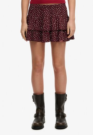 Person trägt einen kurzen, bordeauxfarbenen Blumenrock und dunkelbraune Lederstiefel mit Schnallendetails, die bis zur Mitte der Wade reichen, und steht vor einem schlichten Hintergrund.