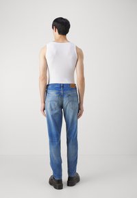 Pánské modré džíny s tapered střihem, s bledým praním a koženou nášivkou na zadním pásu, v kombinaci s bílým tílkem.