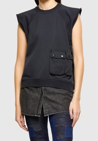 Black sleeveless sweatshirt with ribbed neckline, side pocket featuring snap buttons, and relaxed fit, layered over a dark denim shirt.