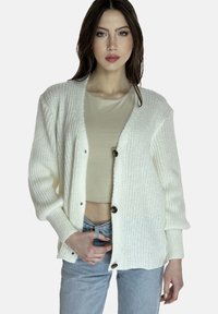 Cárdigan blanco de punto con un corte holgado, que presenta un escote en V profundo, mangas largas y botones marrones. Lleva encima una blusa beige clara y unos vaqueros.