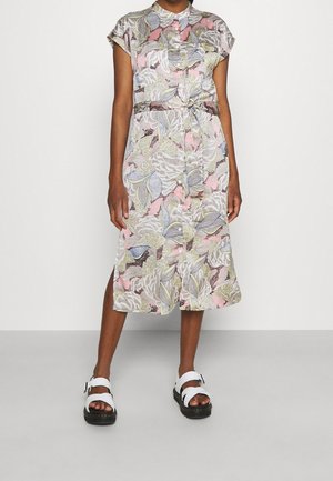 Femme portant une robe fleurie mi-longue boutonnée avec une taille nouée et des sandales blanches à semelles noires.