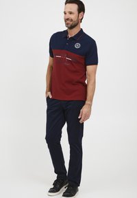 Polo shirt rosso e blu con colletto, presenta un logo e testo grafico sul davanti. Abbina a pantaloni blu navy e sneakers nere.