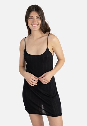 Jeune femme aux longs cheveux foncés portant une robe noire sans manches, souriante et joignant les mains devant un fond clair uni.
