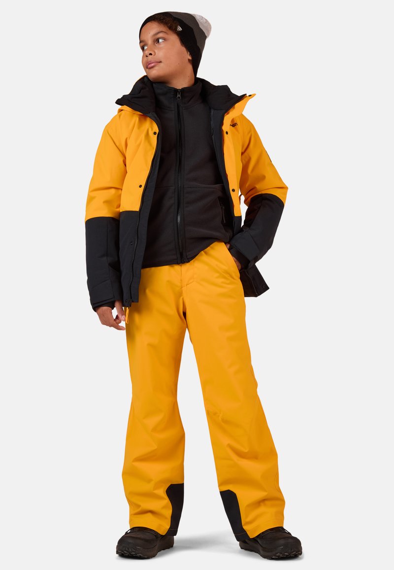 Geel waterdichte jack met zwarte accenten, gecombineerd met bijpassende gele broek. Een zwarte fleece binnenlaag en sneeuwlaarzen maken de outfit compleet.