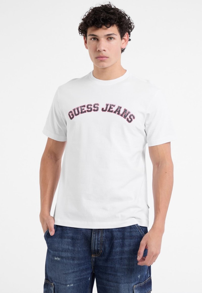 Balta medvilnė T marškiniai su kreiva "GUESS JEANS" logotipu raudonos ir tamsiai mėlynos spalvos siuvinėjimu. Apvalios kaklo iškirptės ir trumpomis rankovėmis. Dėvimi su džinsiniais šortais.