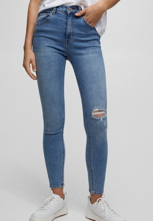 Personne portant un jean skinny bleu clair avec un genou déchiré et des baskets blanches, debout devant un fond uni clair.