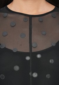 Čierny priehľadný top so zvýraznenými sivými bodkami s textúrovaným povrchom. Výstrih je zaoblený s viditeľným švom uprostred.