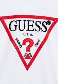 T-shirt bianco con un logo a triangolo rosso contenente il testo "GUESS", un punto interrogativo, e le etichette "WASHED" e "JEANS" in nero.