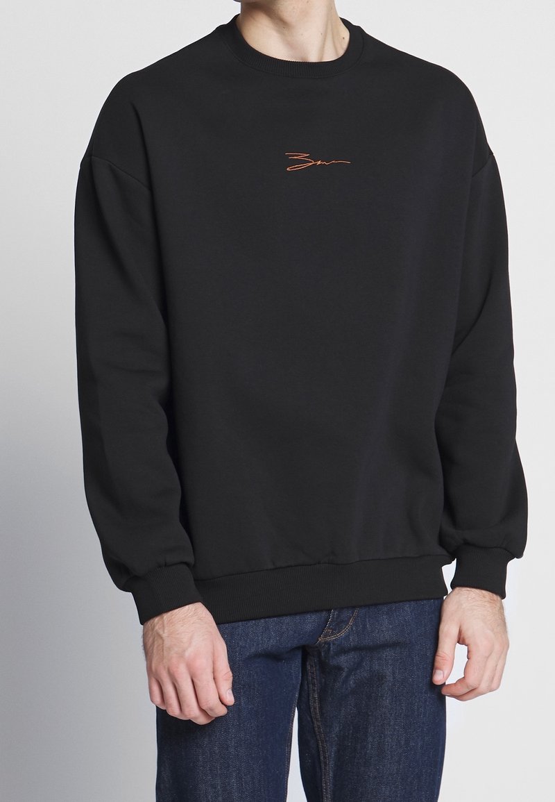 Personne portant un sweat-shirt noir uni à col rond avec un petit détail de signature orange sur la poitrine et un jean bleu sur un fond uni.