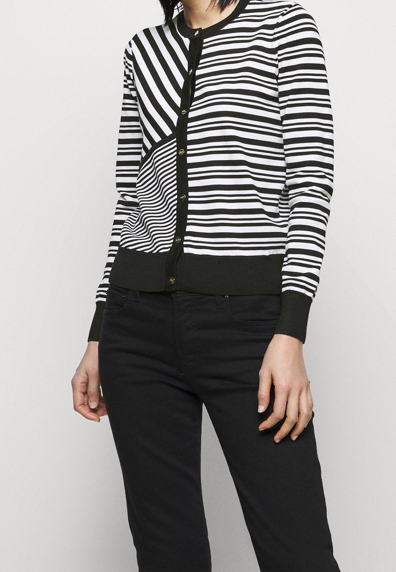 Femme portant un cardigan boutonné à rayures noires et blanches avec des directions de rayures variées et un pantalon noir uni.