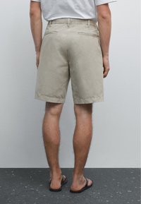 Beige Shorts mit glatter Textur, entspannter Passform und gesäumten Kanten. Zwei Gesäßtaschen und ein Knopfverschluss an der Taille.
