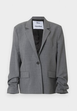 Mladá žena nosiaca sivý oversized blejzer, biely top, svetlomodré džínsy, čierny opasok a čierne vysoké podpätky, stojaca pred jednoduchým pozadím.