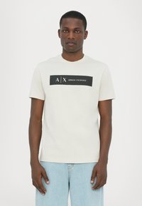 Kortermet, lys beige t-skjorte laget av bomull, med en svart rektangulær grafikk med teksten "A/X ARMANI EXCHANGE" i midten.