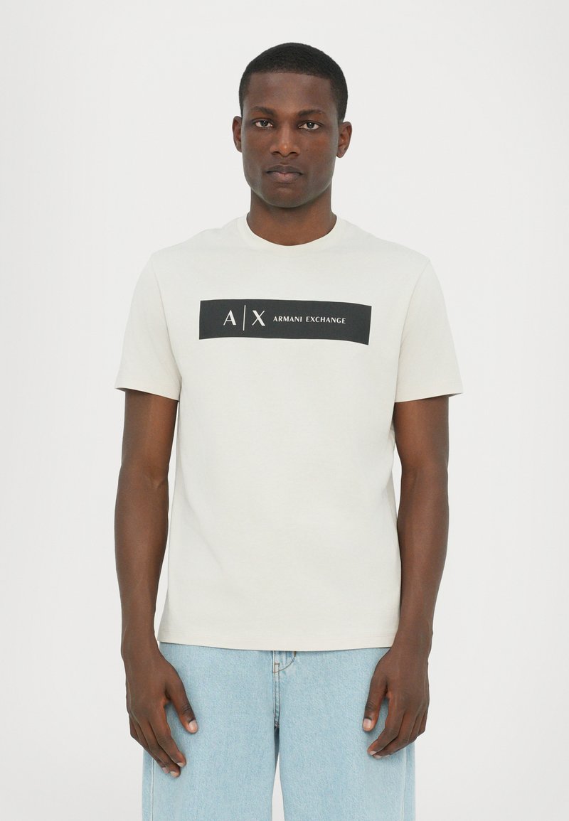 Kortermet, lys beige t-skjorte laget av bomull, med en svart rektangulær grafikk med teksten "A/X ARMANI EXCHANGE" i midten.