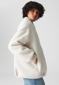 Witte fleece jas met ribbels bij de manchetten en kraag. Oversized pasvorm met zijzakken. Zachte textuur en relaxte ontwerp, gecombineerd met een blauwe spijkerbroek.