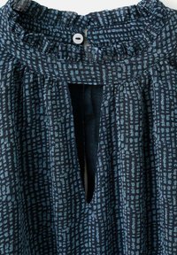 Blusa azul marino con un patrón texturizado de cuadrados azules más claros, con un escote fruncido y una apertura en forma de llave, asegurada por un botón.