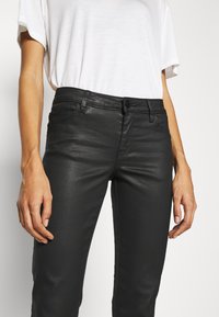 Person som bär åtsittande svarta belagda byxor och en enkel vit t-shirt med kort ärm, stående mot en enkel ljus bakgrund.