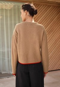 Jasnobrązowy krótki sweter z prążkowanym wykończeniem i czerwonym akcentem, połączony z czarnymi spodniami typu wide-leg. Tkanina wydaje się miękka i teksturowana.