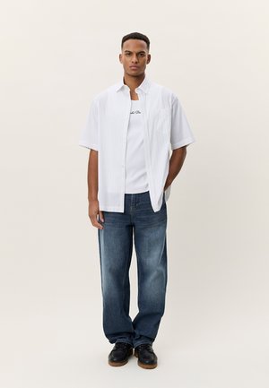 Νεαρός άνδρας στέκεται φορώντας λευκό πουκάμισο με κοντά μανίκια, λευκό μπλουζάκι με κείμενο, μπλε τζιν και μαύρα παπούτσια σε ουδέτερο φόντο.