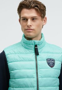 Gilet trapuntato azzurro chiaro con colletto alto, chiusura a zip e un patch con il logo sul petto. Presenta una texture liscia e lucida.