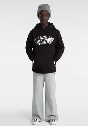 Sort hættetrøje med hvid "VANS OFF THE WALL" grafik, parret med lysegrå sweatpants. Modellen bærer en grå strikket hue og ternede slip-on sko.