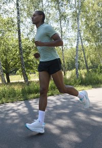 Chemise à manches courtes vert clair, shorts de course noirs, chaussures de sport blanches avec des accents turquoise, courant sur un chemin entouré d'arbres.
