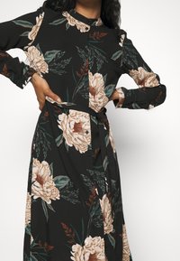 Robe florale noire avec de grandes pivoines beiges, des feuilles vertes et des accents subtils aux tons terre; devant boutonné et détail de ceinture à la taille.