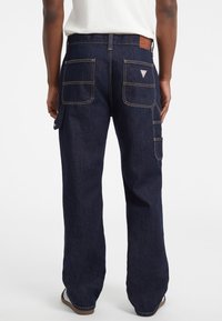 Persona vestida con pantalones cargo de mezclilla azul oscuro con grandes bolsillos traseros y un bolsillo lateral, combinados con zapatos blancos y una camisa blanca.