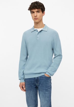 Jeune homme portant un pull polo à manches longues bleu clair et un jean bleu, debout avec une main dans la poche devant un fond uni.
