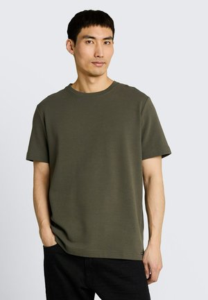 Mężczyzna o krótkich czarnych włosach, ubrany w oliwkową teksturowaną koszulę z krótkim rękawem i czarne spodnie, stojący z jedną ręką w kieszeni.