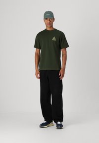 T-shirt en coton vert avec un logo en triangle jaune, associé à un pantalon large noir et des sneakers bleues avec des lacets blancs.