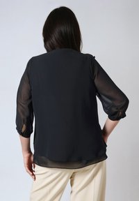 Zwarte doorschijnende blouse met driekwartmouwen, met een vloeiende uitstraling en een licht gebogen zoom. In lichte broek gestopt.
