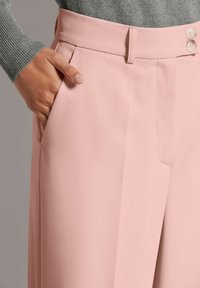 Lichtroze, op maat gemaakte broek met een brede pijp, zijzak en een tailleband met twee knopen. De stof lijkt glad met een lichte glans.