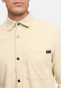 Camisa de pana color crema con cuello, botones negros, un bolsillo en el pecho y una etiqueta en el bolsillo con texto.