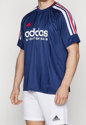 Man draagt een marineblauw Adidas sportshirt met witte, rode en blauwe strepen op de schouders, gecombineerd met witte Adidas shorts.