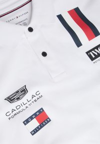 Λευκό πόλο μπλουζάκι Tommy Hilfiger με το λογότυπο της ομάδας Cadillac Formula 1 και ταμπελάκια σε κόκκινο, άσπρο και ναυτικό μπλε.