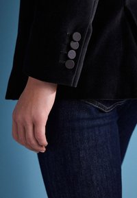 Blazer en velours noir avec une coupe cintrée, doté de quatre boutons ronds assortis sur le poignet et d'un jean en denim bleu en gros plan.