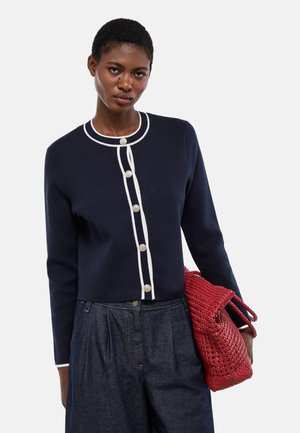 Jeune femme portant un cardigan bleu marine avec des bordures et des boutons blancs, un jean large foncé, tenant un sac à main tressé rouge.