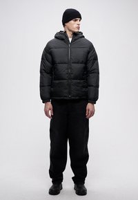 Sort dunjakke med hette, som har quiltede seksjoner, frontglidelås og en avslappet passform, kombinert med svarte bukser og en lue.