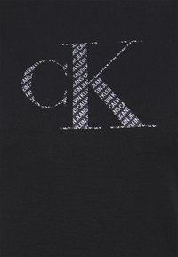 Czarny T-shirt z dużym, białym nadrukiem liter "ck" oraz powtarzającym się tekstem "CALVIN KLEIN JEANS" w mniejszej czcionce.