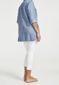 Donna in piedi con il lato destro visibile, che indossa una camicia di jeans azzurro chiaro, pantaloni bianchi tagliati e sandali aperti color beige su uno sfondo bianco.