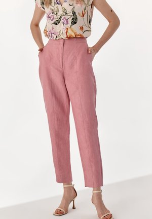 Kvinde iført lyserøde hørbukser med høj talje og forreste sømme, kombineret med blomstrede ærmeløse bluse og beige hælede sandaler med ankelrem.